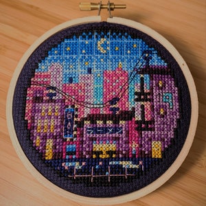 May include: A circular embroidery hoop featuring a colourful cross-stitch design depicting a cityscape at night. The design incorporates a range of colours, including pink, blue, purple, and yellow, and includes a crescent moon and stars in the sky.