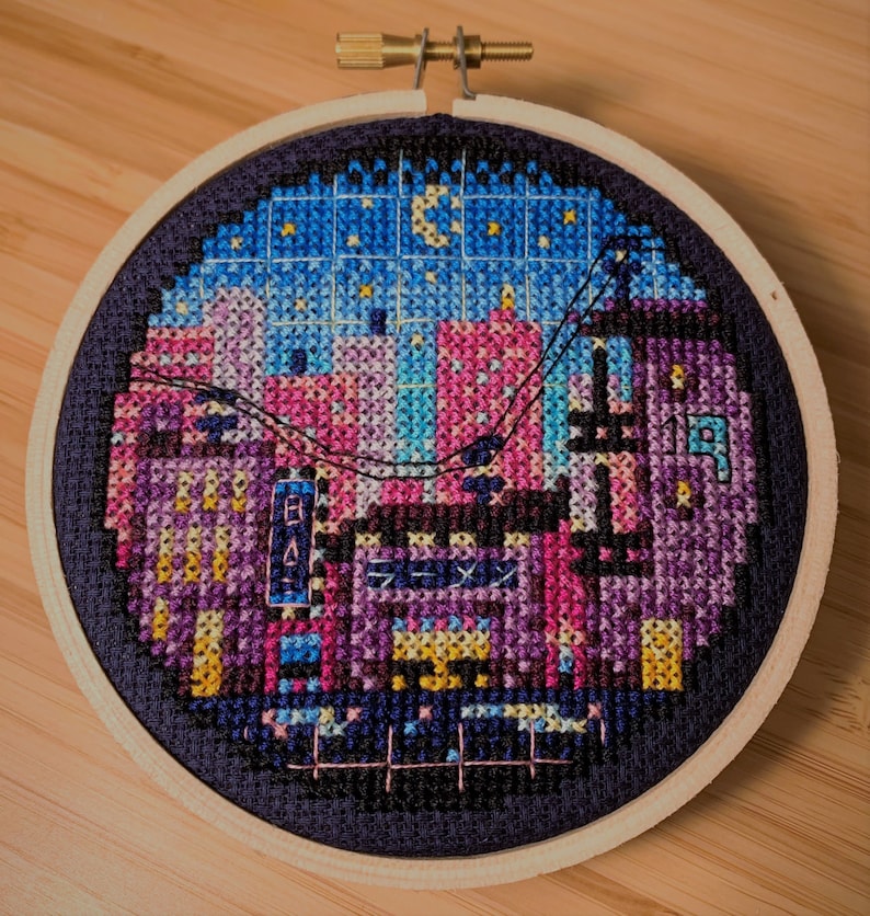 May include: A circular embroidery hoop featuring a colourful cross-stitch design depicting a cityscape at night. The design incorporates a range of colours, including pink, blue, purple, and yellow, and includes a crescent moon and stars in the sky.
