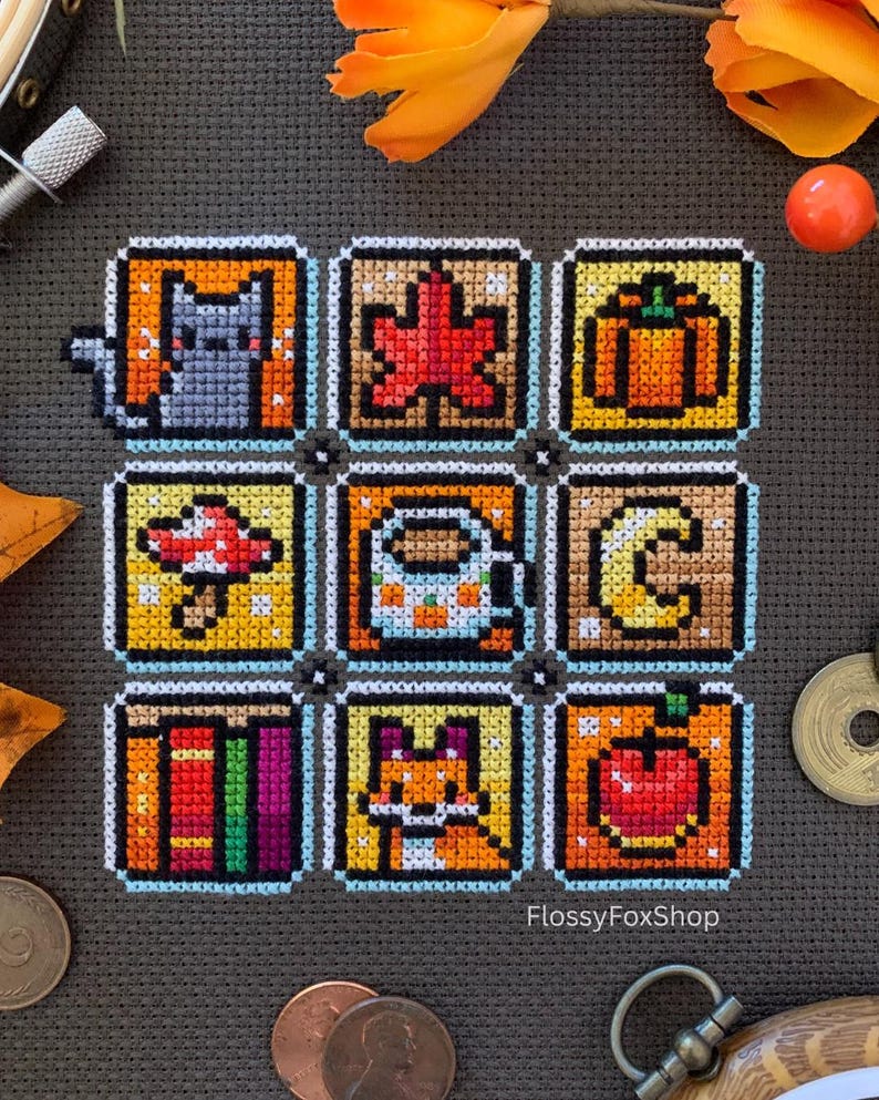 May include: A cross-stitch sampler with nine square designs depicting autumn themes. Designs include a cat, maple leaf, pumpkin, mushroom, coffee mug, crescent moon, books, fox, and apple. The text "FlossyFoxShop" is visible.