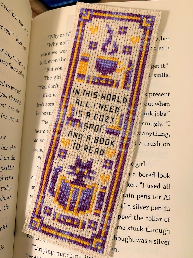 Peut inclure: Marque-page au point de croix avec un fond cr&egrave;me, des motifs g&eacute;om&eacute;triques violets et dor&eacute;s et le texte "IN THIS WORLD ALL I NEED IS A COZY SPOT AND A BOOK TO READ". Le marque-page est rectangulaire et dans un livre ouvert.