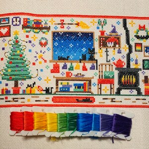 Puede incluir: Un patr&oacute;n de punto de cruz que representa una escena de invierno con un &aacute;rbol de Navidad, una chimenea y un gato. El patr&oacute;n est&aacute; en tela blanca con hilo rojo, verde y azul. El patr&oacute;n est&aacute; enmarcado con un borde rojo.