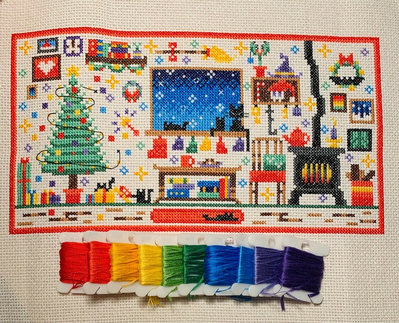 Puede incluir: Un patr&oacute;n de punto de cruz que representa una escena de invierno con un &aacute;rbol de Navidad, una chimenea y un gato. El patr&oacute;n est&aacute; en tela blanca con hilo rojo, verde y azul. El patr&oacute;n est&aacute; enmarcado con un borde rojo.
