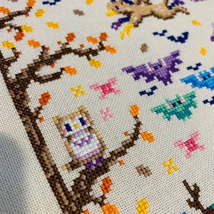 May include: Cross-stitch pattern featuring a brown owl perched on a branch with orange and yellow leaves. The owl is wearing a purple and white dress. There are bats and other flying creatures in the background.
