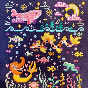 May include: A cross-stitch embroidery design featuring a pink whale with a crown, a crescent moon, clouds, and a variety of fish and sea creatures. The design is set against a dark blue background with white and yellow accents.