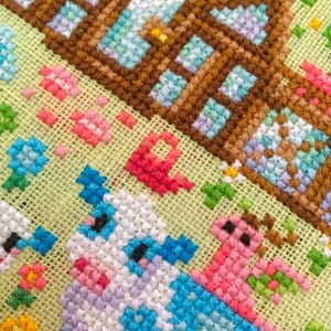 May include: Close-up of a cross-stitch design featuring a brown house with blue windows and a cow with blue and white patches. Pink and red flowers and green foliage are also visible.