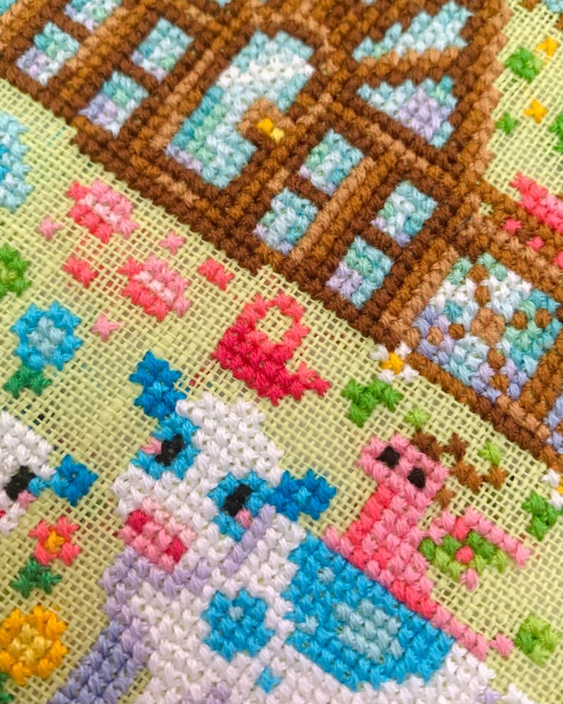 May include: Close-up of a cross-stitch design featuring a brown house with blue windows and a cow with blue and white patches. Pink and red flowers and green foliage are also visible.