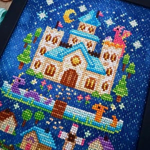 Op de afbeelding: Een kruissteekborduurwerk in een zwarte lijst met een groot kasteel met blauwe daken, een gele maansikkel en sterren tegen een donkerblauwe achtergrond. Onder het kasteel staan kleine huisjes, een hek en twee draken in een blauwe rivier.