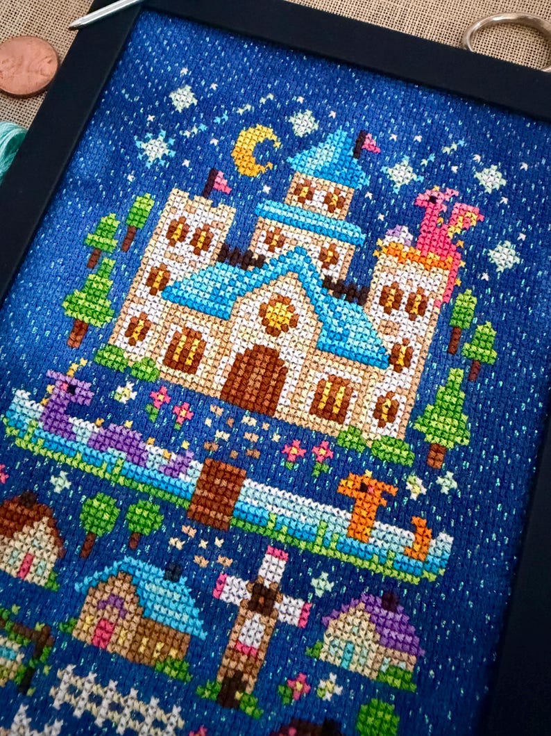 Op de afbeelding: Een kruissteekborduurwerk in een zwarte lijst met een groot kasteel met blauwe daken, een gele maansikkel en sterren tegen een donkerblauwe achtergrond. Onder het kasteel staan kleine huisjes, een hek en twee draken in een blauwe rivier.