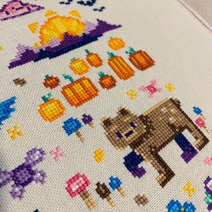 May include: Cross-stitch pattern featuring a brown bear, pumpkins, mushrooms, and a purple sunset. The pattern is stitched on a white fabric.