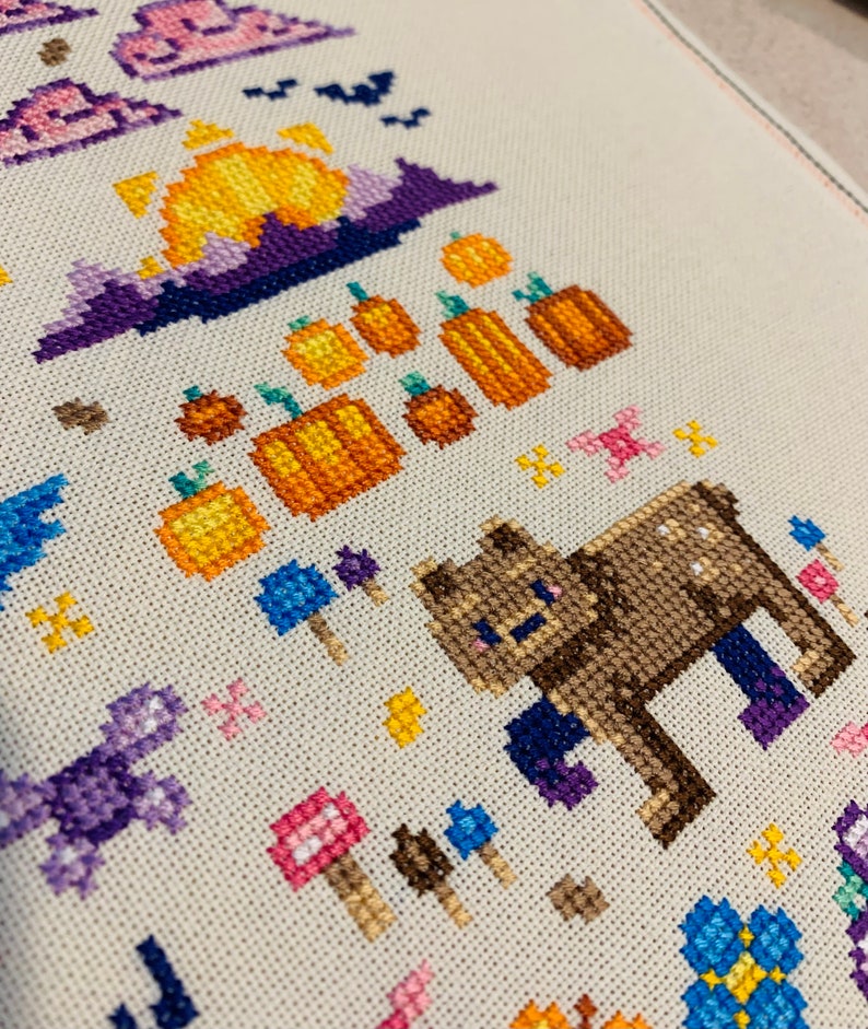 May include: Cross-stitch pattern featuring a brown bear, pumpkins, mushrooms, and a purple sunset. The pattern is stitched on a white fabric.