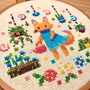 May include: A cross-stitch embroidery hoop depicting a fox wearing a blue dress and holding a watering can, surrounded by flowers and plants. The embroidery is crafted in a range of colours, including pink, blue, green, and yellow.