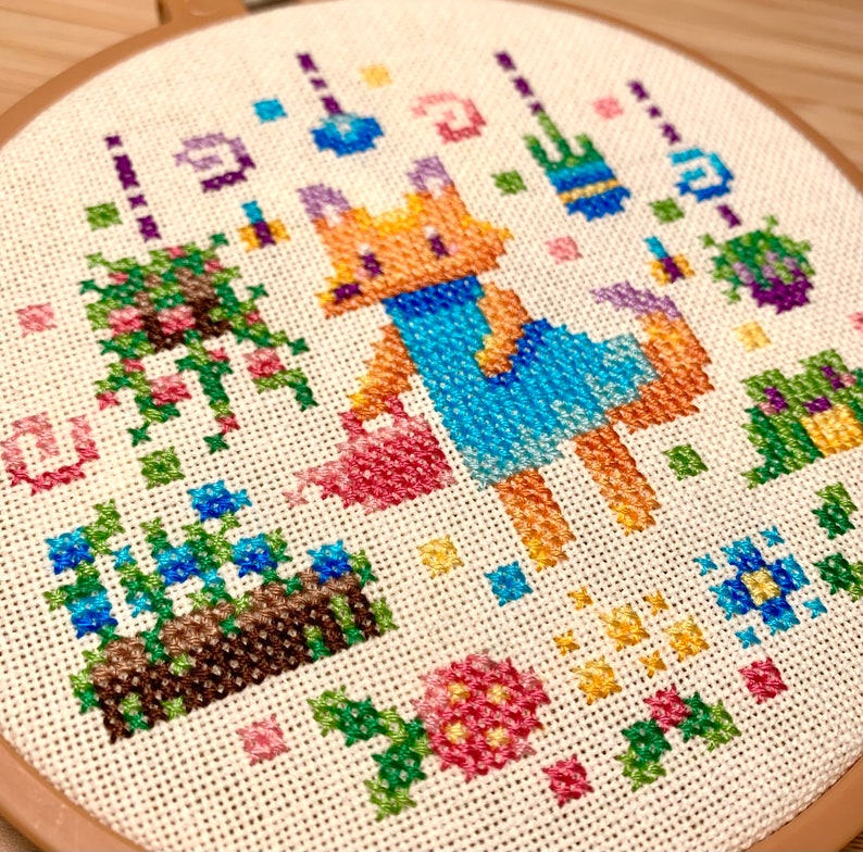 May include: A cross-stitch embroidery hoop depicting a fox wearing a blue dress and holding a watering can, surrounded by flowers and plants. The embroidery is crafted in a range of colours, including pink, blue, green, and yellow.
