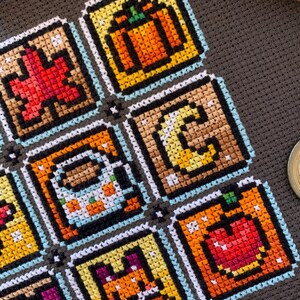 May include: A close-up of a cross-stitch sampler with pixel art designs, including a pumpkin, apple, fox, and coffee mug. The sampler is stitched with various colours and features autumn-themed icons.