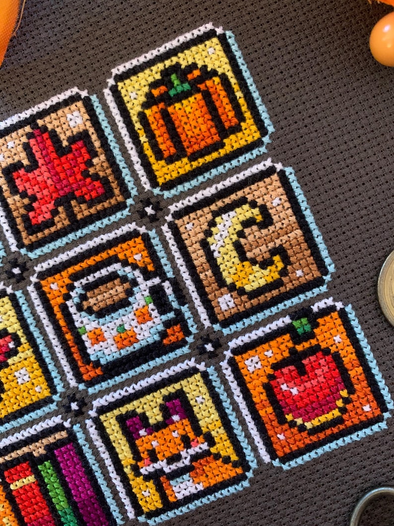 May include: A close-up of a cross-stitch sampler with pixel art designs, including a pumpkin, apple, fox, and coffee mug. The sampler is stitched with various colours and features autumn-themed icons.