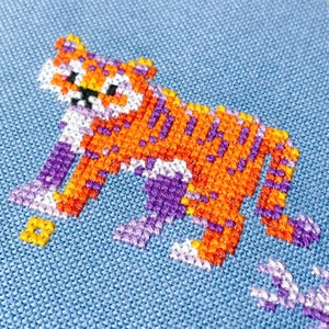 May include: A cross-stitch embroidery of a tiger with orange, purple, and white thread on a blue fabric background.