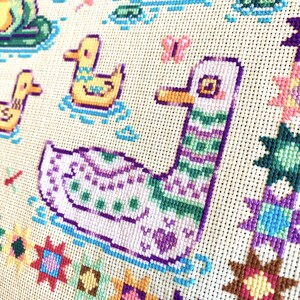 May include: Cross-stitch embroidery featuring a purple and green duck swimming in a blue pond. The duck is surrounded by colorful flowers and stars.
