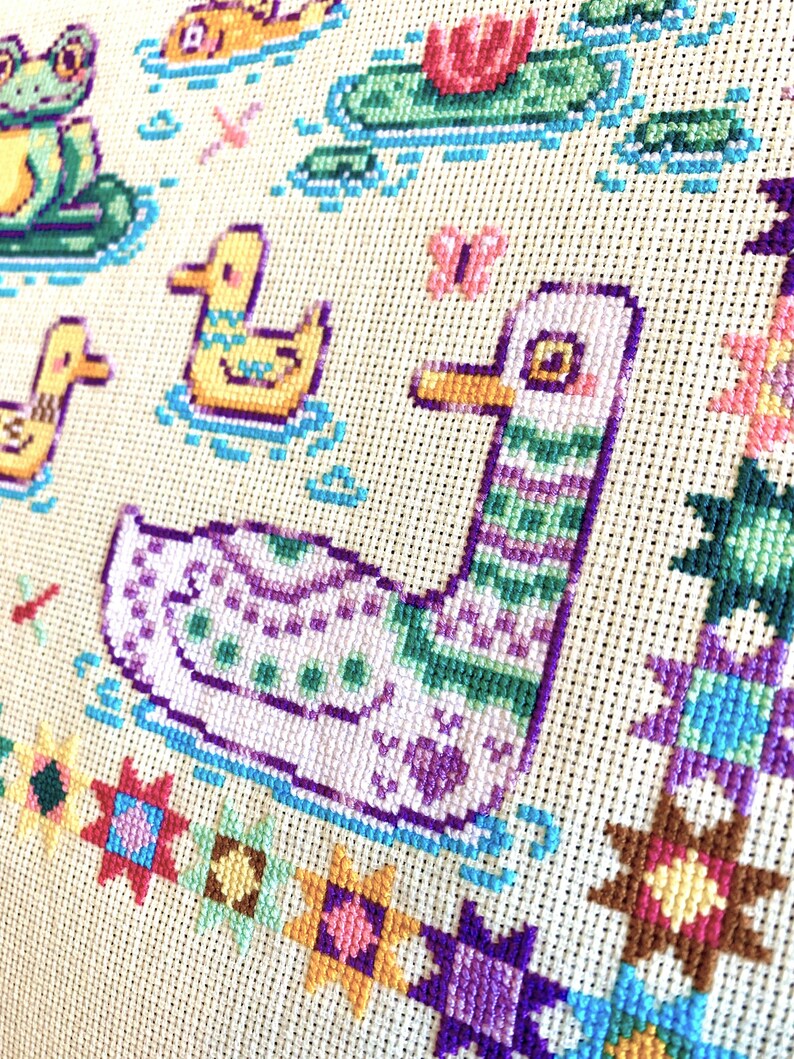 May include: Cross-stitch embroidery featuring a purple and green duck swimming in a blue pond. The duck is surrounded by colorful flowers and stars.