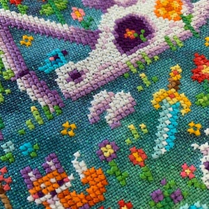 Puede incluir: Primer plano de un dise&ntilde;o de punto de cruz que presenta un unicornio blanco con detalles morados y naranjas, rodeado de flores coloridas y un fondo azul. La labor de aguja es detallada y vibrante, mostrando varias puntadas.