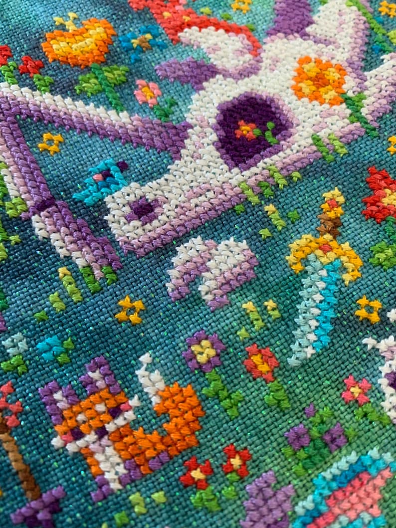 Puede incluir: Primer plano de un dise&ntilde;o de punto de cruz que presenta un unicornio blanco con detalles morados y naranjas, rodeado de flores coloridas y un fondo azul. La labor de aguja es detallada y vibrante, mostrando varias puntadas.