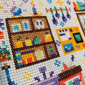 May include: A colorful cross-stitch pattern depicting a room with a bookshelf, a window, and a hanging mobile. The bookshelf is filled with books and other items, and the window is open to reveal a view of a garden.
