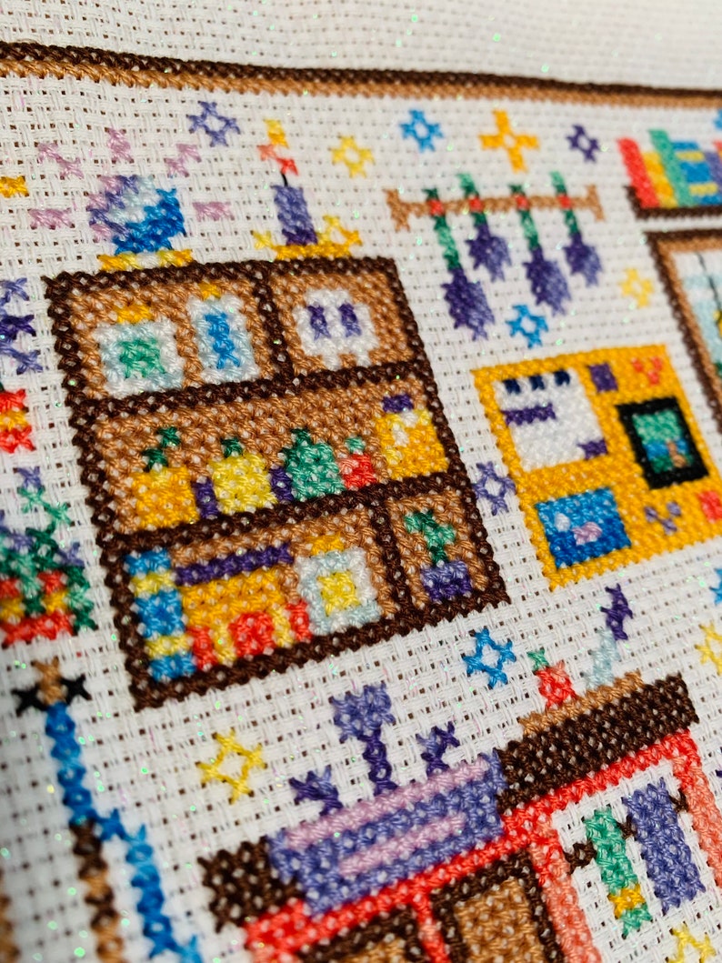 May include: A colorful cross-stitch pattern depicting a room with a bookshelf, a window, and a hanging mobile. The bookshelf is filled with books and other items, and the window is open to reveal a view of a garden.