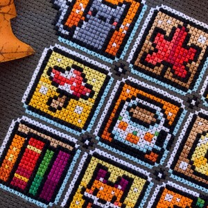 May include: A handmade cross-stitch piece with nine square pixel art designs. The images include a cat, a maple leaf, a mushroom, a coffee cup, a stack of books, a fox, and an apple. The squares are outlined in black and white, with a light blue border.