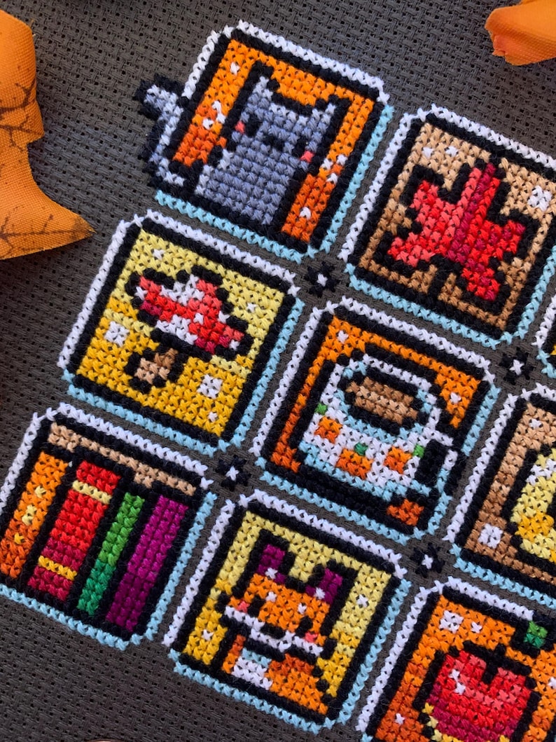 May include: A handmade cross-stitch piece with nine square pixel art designs. The images include a cat, a maple leaf, a mushroom, a coffee cup, a stack of books, a fox, and an apple. The squares are outlined in black and white, with a light blue border.