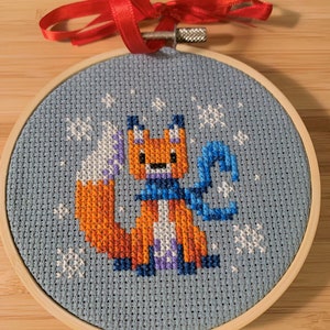 May include: A blue and white cross-stitch ornament featuring a fox wearing a blue scarf and standing in a snowy scene. The ornament is framed in a wooden hoop.