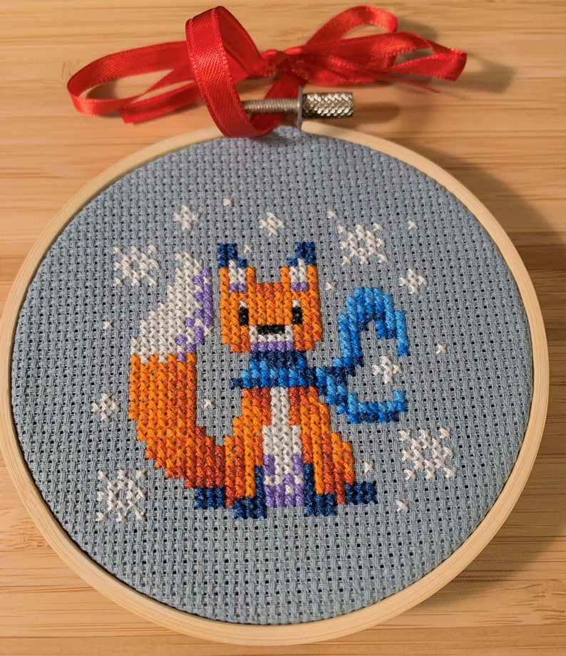May include: A blue and white cross-stitch ornament featuring a fox wearing a blue scarf and standing in a snowy scene. The ornament is framed in a wooden hoop.
