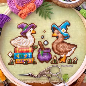 May include: Cross-stitch embroidery featuring two ducks wearing witch hats, with a purple cauldron and flowers. The ducks are in a hoop with embroidery floss and a pair of decorative scissors. A small crocheted hat is at the top.