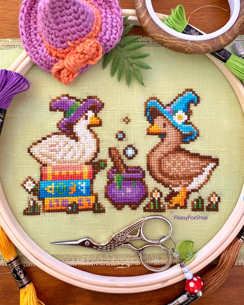 May include: Cross-stitch embroidery of two ducks in witch hats, with a purple cauldron and flowers. The ducks are in a hoop with embroidery floss and decorative scissors. A small crocheted hat is at the top.