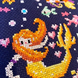 May include: A cross-stitch pattern of a mermaid with a yellow tail and orange hair on a dark blue background. The mermaid is surrounded by small white, pink, and blue flowers.