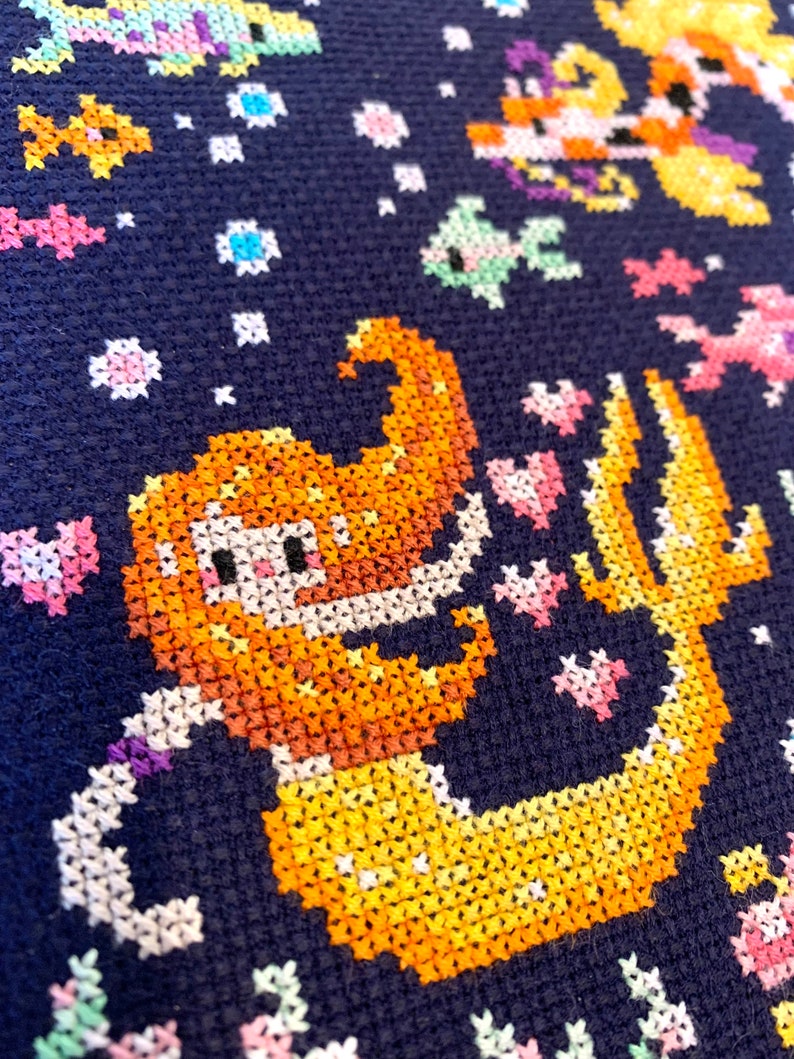 May include: A cross-stitch pattern of a mermaid with a yellow tail and orange hair on a dark blue background. The mermaid is surrounded by small white, pink, and blue flowers.