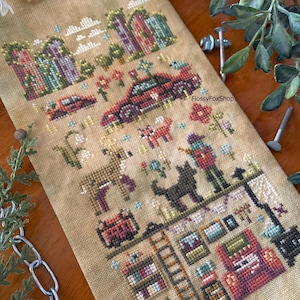 May include: Cross-stitch sampler depicting a rural scene with houses, a car, animals, and a person. The design includes a deer, a black cat, and a person with a hat. The color palette includes shades of brown, green, red, and purple.