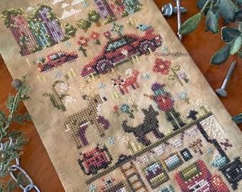 May include: Cross-stitch sampler featuring a rural scene with houses, a car, animals, and a person. The design includes a deer, a black cat, and a person with a hat. The color palette includes shades of brown, green, red, and purple.