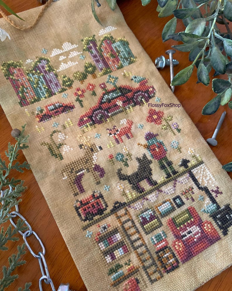 May include: Cross-stitch sampler featuring a rural scene with houses, a car, animals, and a person. The design includes a deer, a black cat, and a person with a hat. The color palette includes shades of brown, green, red, and purple.