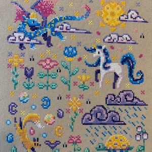 Puede incluir: Un bordado de punto de cruz con un drag&oacute;n azul y morado, un unicornio blanco, un sol amarillo, nubes, flores, un zorro y una tortuga. El bordado est&aacute; hecho en una tela beige.