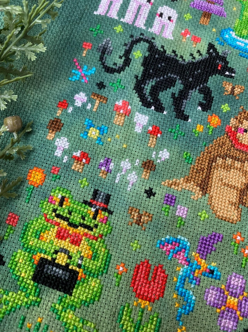 May include: A green cross-stitch embroidery featuring a variety of whimsical creatures, including a frog wearing a top hat, a dragon, a bear, and a black monster. The background is a green field with flowers and mushrooms.