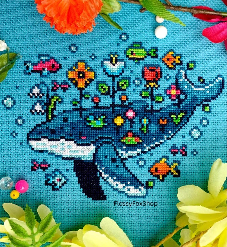 Puede incluir: Arte de punto de cruz que representa una ballena azul adornada con flores y peces. La ballena es de color azul, con detalles blancos. Flores y peces coloridos rodean la ballena sobre un fondo azul claro. El texto "FlossyFoxShop" es visible.
