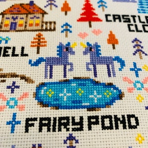 May include: Cross-stitch design featuring a fairy tale scene with a house, trees, unicorns, and a pond. The words "Fairy Pond" and "Castle Cloud" are stitched in black. The design uses various colors including blue, orange, and pink.