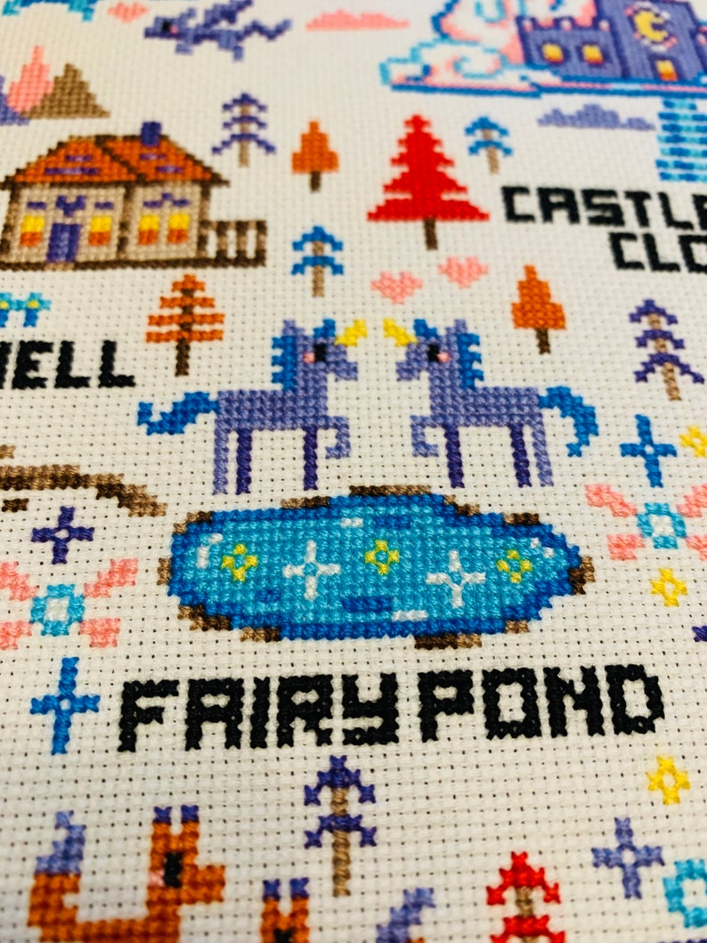 May include: Cross-stitch design featuring a fairy tale scene with a house, trees, unicorns, and a pond. The words "Fairy Pond" and "Castle Cloud" are stitched in black. The design uses various colors including blue, orange, and pink.
