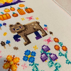 May include: Cross-stitch embroidery featuring a brown bear with purple boots, surrounded by orange pumpkins, blue flowers, and purple bells. The design is on a white fabric background.
