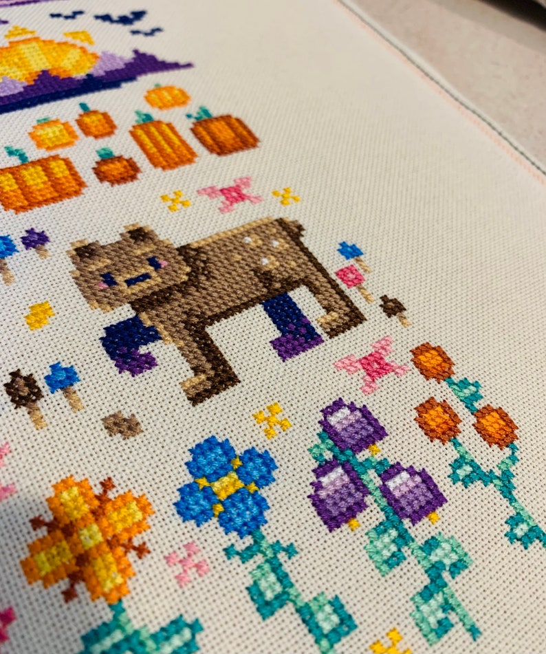 May include: Cross-stitch embroidery featuring a brown bear with purple boots, surrounded by orange pumpkins, blue flowers, and purple bells. The design is on a white fabric background.