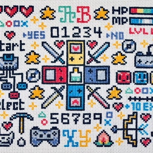 Puede incluir: Bordado a punto de cruz con elementos de videojuegos pixelados. Incluye corazones, estrellas, espadas, mandos y texto como "Start" y números. La paleta de colores es vibrante, con rojos, azules, amarillos y verdes.
