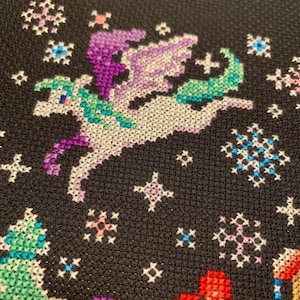 May include: Cross-stitch embroidery on black fabric depicting a white unicorn with purple and green wings flying through a starry night sky. The unicorn is surrounded by stars, snowflakes, and flowers in various colors.