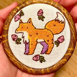 May include: A cross-stitch embroidery of an orange fox with purple legs standing on a white background surrounded by pink flowers with green leaves. The embroidery is framed in a round wooden frame.