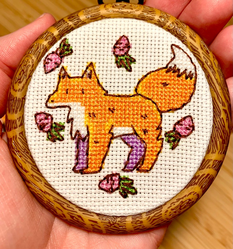 May include: A cross-stitch embroidery of an orange fox with purple legs standing on a white background surrounded by pink flowers with green leaves. The embroidery is framed in a round wooden frame.