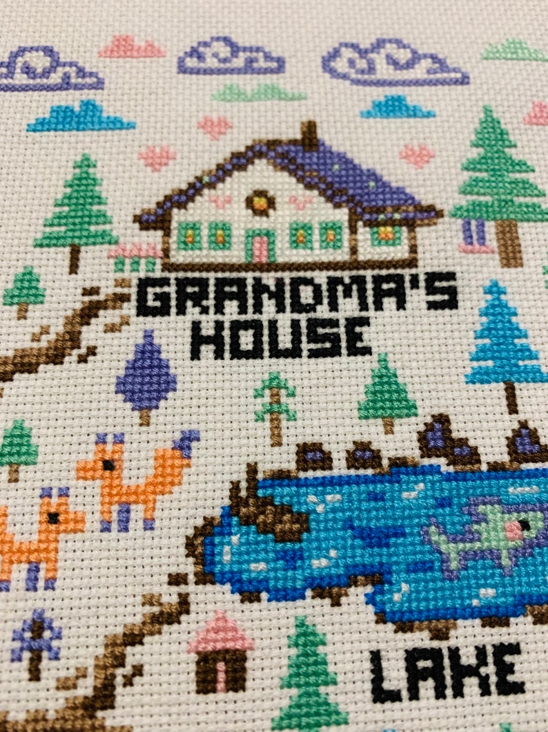 Puede incluir: Bordado de punto de cruz de una casa con el texto "Grandma's House" y un lago con el texto "Lake". La escena incluye &aacute;rboles, nubes, corazones y zorros.