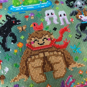 May include: A brown and red cross-stitch pattern of a friendly-looking creature with a red scarf and a red bird on its head. The creature is surrounded by a green background with flowers, mushrooms, and other creatures.