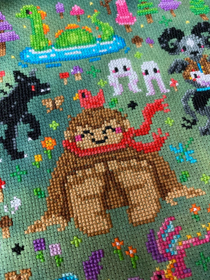 May include: A brown and red cross-stitch pattern of a friendly-looking creature with a red scarf and a red bird on its head. The creature is surrounded by a green background with flowers, mushrooms, and other creatures.